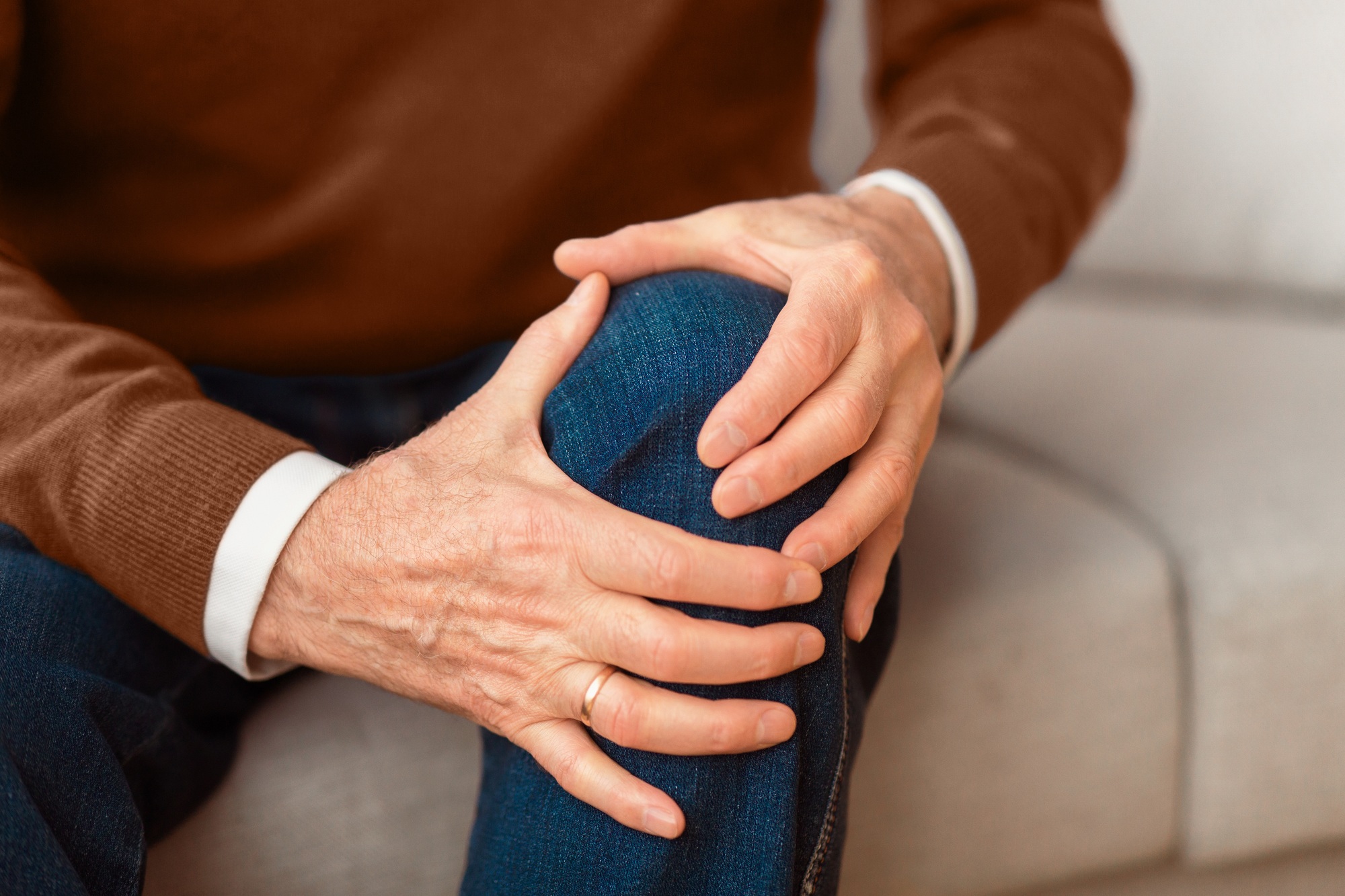 Unrecognizable elderly man massaging knee sitting on couch at home